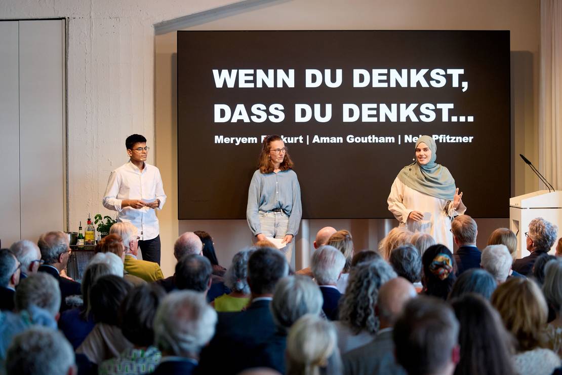 Foto: Drei junge Leute, zwei Frauen und ein Mann, stehen auf einer Bühne, davor ist das Publikum verschwommen zu sehen. Auf die Wand wird projiziert 