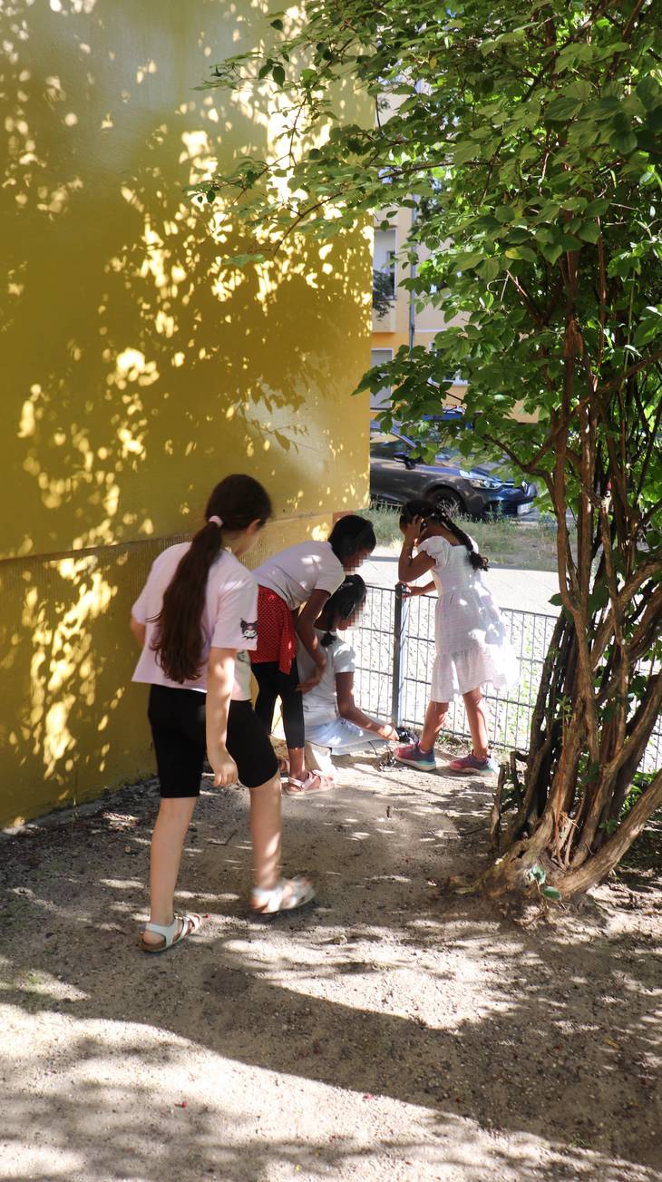 Kinder nehmen an einer gelben Hauswand im Schatten eines Baumes an einem Zaun mit Mikrofonen Umgebungsgeräusche auf